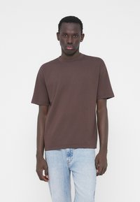 Rjava bombažna majica z okroglim izrezom in kratkimi rokavi, kombinirana s svetlo modrimi jeans hlačami. Preprost dizajn, brez vidnih vzorcev ali poudarkov.