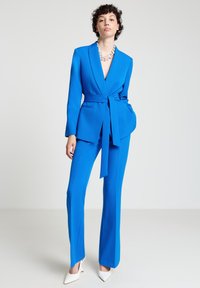 Woman wearing a bright blue belted blazer and matching trousers with white pointed heels, standing against a plain light background.
