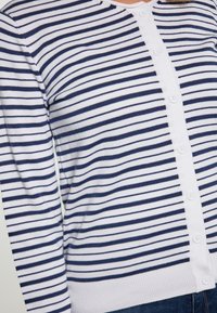Pruhovaný cardigan v bílé a námořnické modré; pletený materiál s knoflíkovým zapínáním a kulatým výstřihem. Vybaven dlouhými rukávy a ribkovanými manžetami.