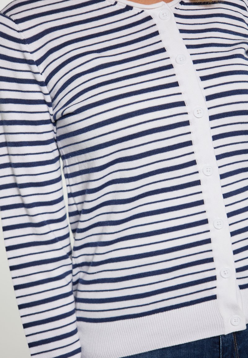 Pruhovaný cardigan v bílé a námořnické modré; pletený materiál s knoflíkovým zapínáním a kulatým výstřihem. Vybaven dlouhými rukávy a ribkovanými manžetami.