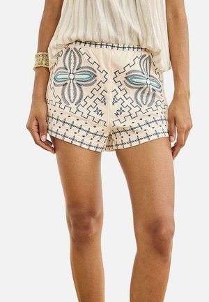 Mujer que lleva pantalones cortos beige con patrones bordados florales y geométricos en azul y blanco, combinados con un top sin mangas a juego.