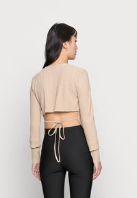 Top de manga longa cropped bege com textura plissada e costas abertas. Tecido liso amarrado na cintura. Calças pretas ajustadas visíveis.