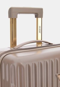 Hard-shell suitcase in light beige with vertical ribbed texture, featuring a gold metal telescopic handle and branded accents.