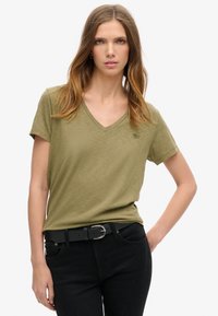 Oliven grøn V-hals t-shirt lavet af blødt stof, med korte ærmer og en subtil logo detalje. Båret med sorte jeans og et sort bælte.