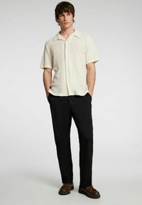 Koszula z krótkim rękawem w jasnym kremowym kolorze, wykonana z dzianiny, z kołnierzykiem typu spread i delikatną teksturą, w połączeniu z czarnymi spodniami i brązowymi butami.