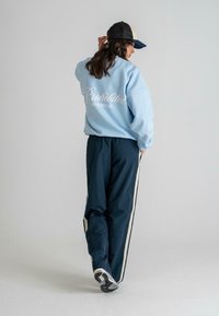 Hellblaues oversized Sweatshirt mit weißem Text auf der Rückseite, kombiniert mit dunkelblauen schmalen Hosen mit cremefarbenen seitlichen Streifen. Schwarze Kappe rückwärts getragen.