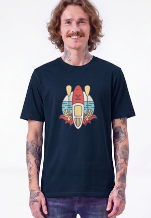 Homme aux longs cheveux et tatouages portant un t-shirt bleu marine avec un motif graphique coloré de kayak et pagaies à l'avant.