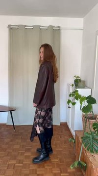 Blazer de couro castanho sobre um vestido de renda preto com fendas. Botas pretas até ao joelho. Cortinas de tom claro ao fundo. Elementos de decoração naturais.