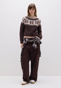 Bruine gebreide trui met een licht bloemenpatroon, gecombineerd met losse bruine cargobroek en een geruit hem dat om de taille is geknoopt. Witte sneakers.