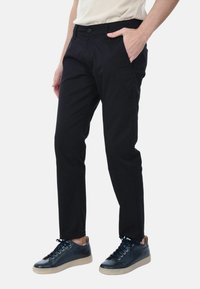 Pantaloni neri in cotone con vestibilità slim, dotati di tasche frontali e chiusura a bottone, abbinati a sneaker navy e una camicia beige.