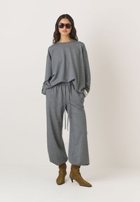 Pilkas laisvalaikio komplektas su laisvu megztiniu ir plataus kirpimo kelnėmis. Minkšta medžiaga, su raišteliu reguliuojama liemens juosta, derinama su rudais smailiais batais ir saulės akinių. 