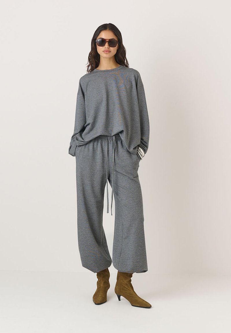 Pilkas laisvalaikio komplektas su laisvu megztiniu ir plataus kirpimo kelnėmis. Minkšta medžiaga, su raišteliu reguliuojama liemens juosta, derinama su rudais smailiais batais ir saulės akinių. 