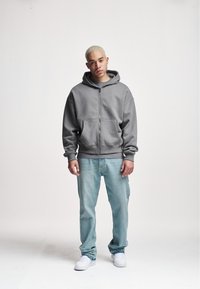Szara bluza na zamek z przestronnym kapturem i przednimi kieszeniami, zestawiona z jasnoniebieskimi dżinsami i białymi tenisówkami na neutralnym tle.