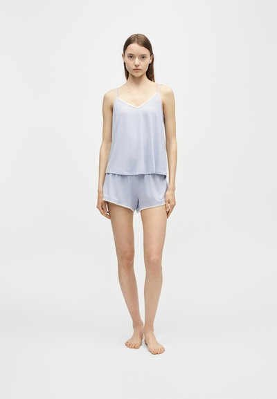Jeune femme debout, pieds nus, portant un ensemble pyjama en satin bleu clair composé d’un caraco et d’un short, sur un fond uni.