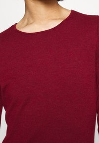 Punainen neulepaita, jossa on pyöreä kaula-aukko ja pitkät hihat, valmistettu pehmeästä kankaasta, jolla on sileä pinta. Ei näkyviä kuvioita tai koristeita.