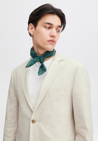 Beige linblazer med en enkel knapp, matchad med en grön mönstrad scarf knuten runt halsen, bärs över en vit skjorta.
