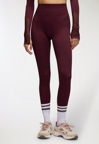 Leggings bordeaux a vita alta con una texture liscia, abbinati a scarpe da ginnastica chiare con accenti beige e rosa e calze bianche a righe.