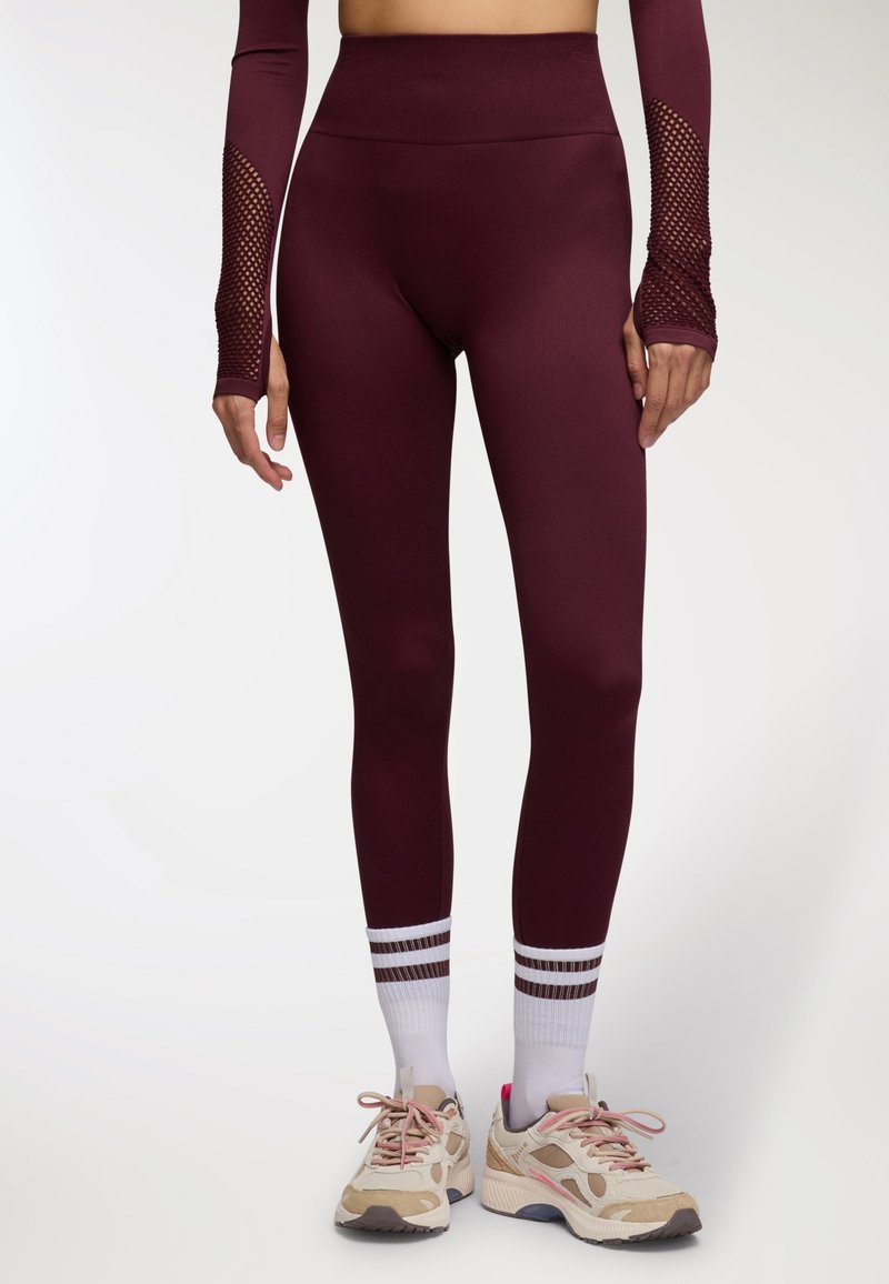 Leggings bordeaux a vita alta con una texture liscia, abbinati a scarpe da ginnastica chiare con accenti beige e rosa e calze bianche a righe.