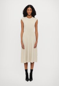 Knit midi-mekko vaaleassa beigesävyssä, joka on varustettu ribbipinnalla, hihattomilla hihoilla ja muotoilulla, joka levenee vyötäröltä. Mustat nilkkurit, joissa on koristeelliset kärkiosat.