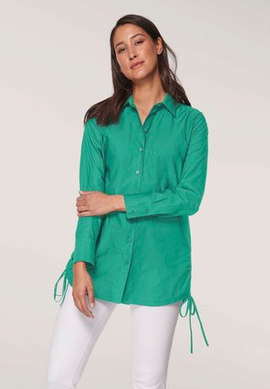 Grünes, knopfbares Hemd mit Kragen, langen Ärmeln und seitlichen Zugkordel-Akzenten. Aus glattem Stoff gefertigt, kombiniert mit weißen Hosen.