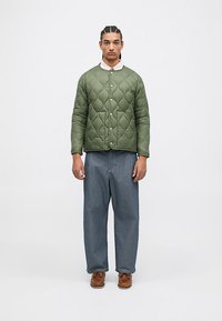 Bărbat cu părul împletit, purtând o geacă verde matlasată, cămașă albă, blugi largi albaștri și pantofi din piele maronie, stând în fața unui fundal simplu.