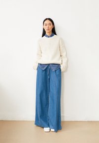 Cremfarbener Strickpullover mit gerippten Bündchen, der über einem blauen Denimhemd getragen wird. Lässige, weit geschnittene blaue Jeanshose und weiße Schuhe.
