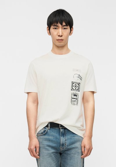 Κρεμ t-shirt με κοντά μανίκια, με μαύρα γραφικά εκτυπωμένα στην αριστερή πλευρά και στρογγυλή λαιμόκοψη. Φορεμένο με μπλε τζιν.