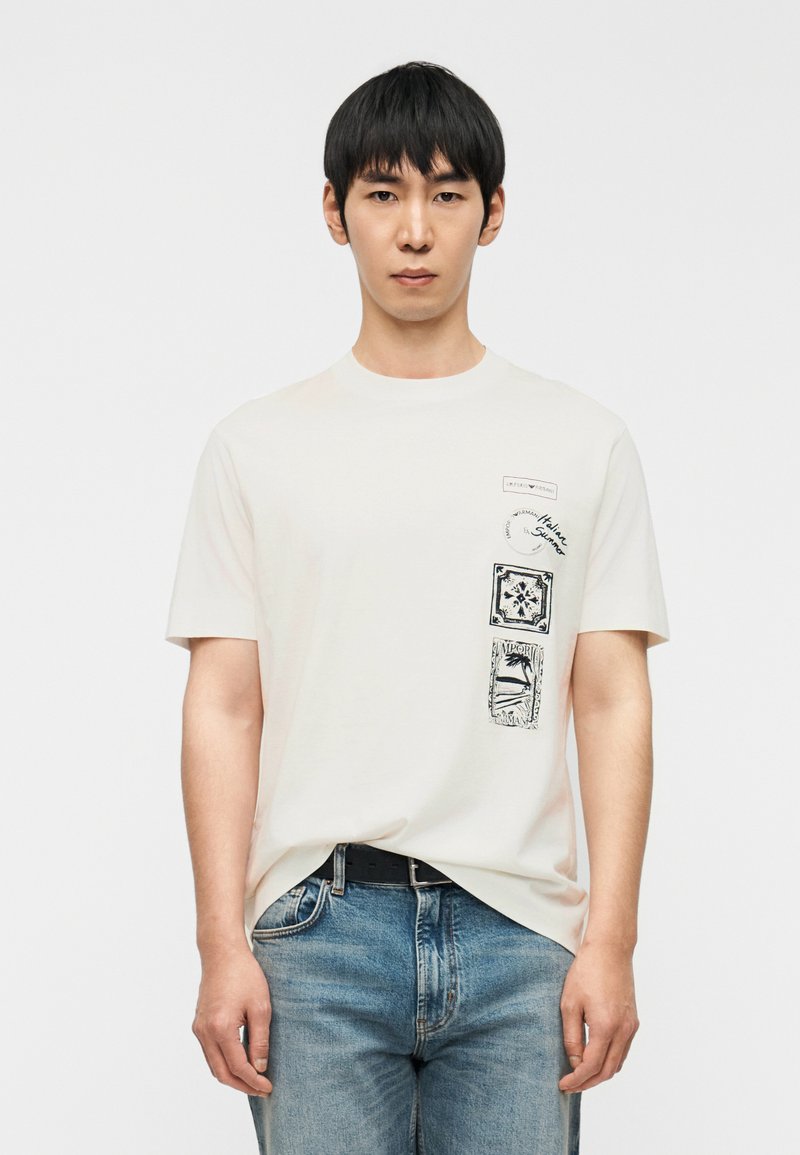 Κρεμ t-shirt με κοντά μανίκια, με μαύρα γραφικά εκτυ πωμένα στην αριστερή πλευρά και στρογγυλή λαιμόκοψη. Φορεμένο με μπλε τζιν.