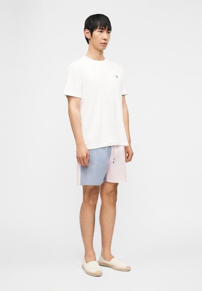 Νέος άνδρας στέκεται φορώντας λευκό κοντομάνικο που κάμισο, σορτς σε μπλε και ροζ μπλοκ χρωμάτων, και μπεζ παπούτσια τύπου slip-on, σε λευκό φόντο.