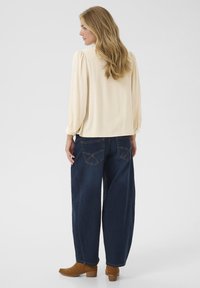Beige strukturierte Bluse mit puffärmeln, kombiniert mit lockeren, dunkelblauen Jeans mit Taschen-Details und braunen Stiefeletten.