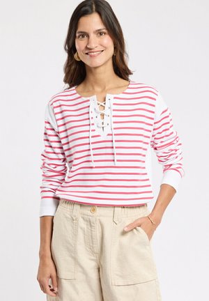 Femme portant une chemise à manches longues rayée blanche et rose avec un col à lacets, un pantalon beige taille haute, debout avec une main dans la poche.