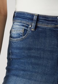 Vaqueros de mezclilla con un corte ajustado, de color azul oscuro, con sutiles detalles desgastados y costuras amarillas. Incluye un bolsillo delantero y cierre de botón metálico.