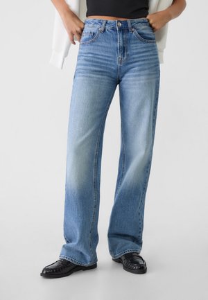 Jean bleu clair taille haute à jambes larges porté avec des mocassins noirs et un crop top noir, les mains reposant dans les poches avant.