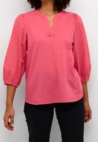 Donna che indossa una blusa texturizzata di colore corallo rosa con maniche a sbuffo a tre quarti e pantaloni neri, in piedi davanti a una parete chiara e uniforme.
