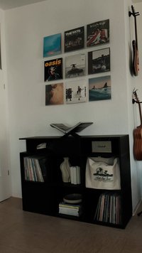 Czarna jednostka regałowa z płytami winylowymi, falistą ceramiczną wazonem, głośnikiem i torbą. Dwie gitary zamocowane na ścianie powyżej.