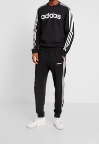 Sweat-shirt noir Adidas avec logo blanc et trois rayures blanches sur les manches, associé à un pantalon de jogging assorti et des baskets blanches.