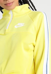 Gul Nike halvdraget sweatshirt med hög krage, vitt logga och marinblå randiga detaljer längs ärmarna. Mjuk, lätt strukturerad tyg.