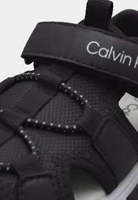 Μαύρο πέδιλο Calvin Klein με ρυθμιζόμενο ιμάντα Velcro και πλεκτά ελαστικά κορδόνια πάνω σε υφασμάτινο επάνω μέρος με υφή και άσπρη άκρη σόλας.