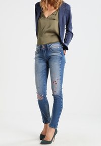 Skinny jeans in light blue denim with floral embroidery detail on knees; paired with an olive silk top and navy blazer. Dark green heels.