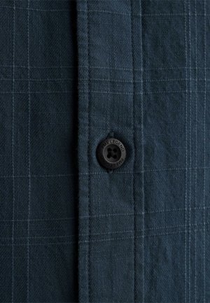 Zbliżenie na ciemnoszarą tkaninę w kratę z czarnym guzikiem z wygrawerowanym napisem "PME LEGEND" przyszytym do listwy koszuli.