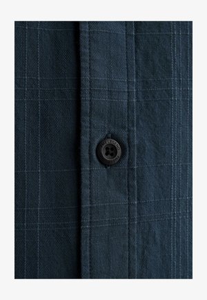 Zbliżenie na ciemnoszarą tkaninę w kratę z czarnym guzikiem z wygrawerowanym napisem "PME LEGEND" przyszytym do listwy koszuli.