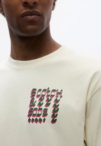 Φούτερ σε κρεμ απόχρωση με πολύχρωμο γραφικό σχέδιο, πράσινο και ροζ κείμενο που γράφει "scotch & soda" και στυλιζαρισμένα φυτικά στοιχεία.