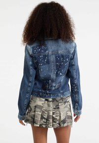 Giacca di jeans blu scuro con motivi di spruzzi più chiari, caratterizzata da un design aderente, chiusura con bottoni e maniche lunghe. Abbinata a una gonna a pieghe in camouflage.