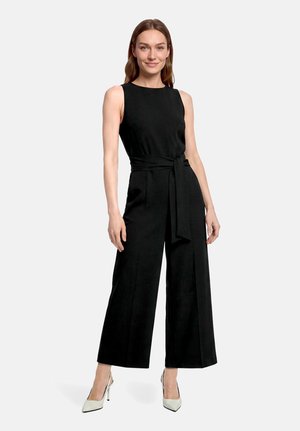 Frau trägt einen ärmellosen schwarzen Jumpsuit mit gebundenem Taillengürtel und weiten Hosen, kombiniert mit weißen High Heels.