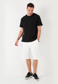 Tricou negru cu mânecă scurtă și buzunar frontal, asociat cu pantaloni scurți albi până la genunchi și adidași negri cu tălpi albe contrastante.