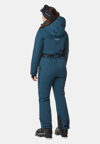 Türkisfarbener Skianzug mit Kapuze, verstellbarem Taillenband, schwarzen Akzenten und Reißverschlusstaschen. Getragen mit Skibrille und schwarzen Skistiefeln.