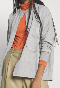 Person med lång flätad hår, orange texturerad polokrage, ljusgrå knappad jacka med logo och högmidjade beige byxor framför vit bakgrund.