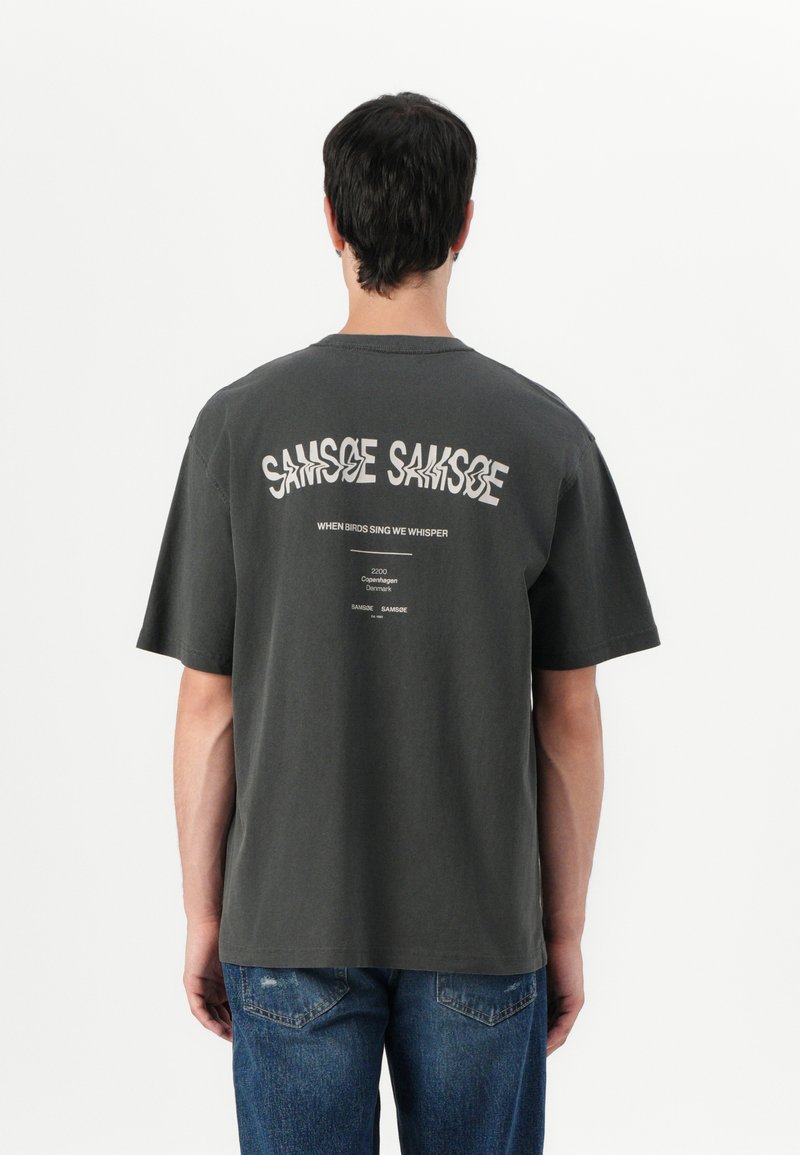 Donkergrijze katoenen t-shirt met een ronde hals. De achterkant heeft een opvallende, vervormde tekstafbeelding en extra kleine tekst. Korte mouwen.