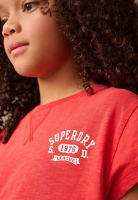 Röd bomulls-T-shirt med korta ärmar och rund halsringning. Framsidan har en grafisk text som visar "SUPERDRY 1978 LEAGUE" i vit, strukturerad lettering.