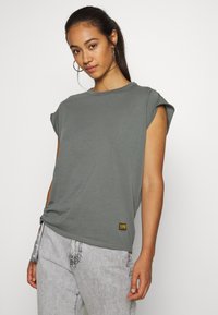 Tricou scurt gri, cu mâneci scurte și guler rotund, dintr-un material moale, având un detaliu cu nod pe lateral și o etichetă galbenă la tiv. Purtați-l cu blugi gri deschis.